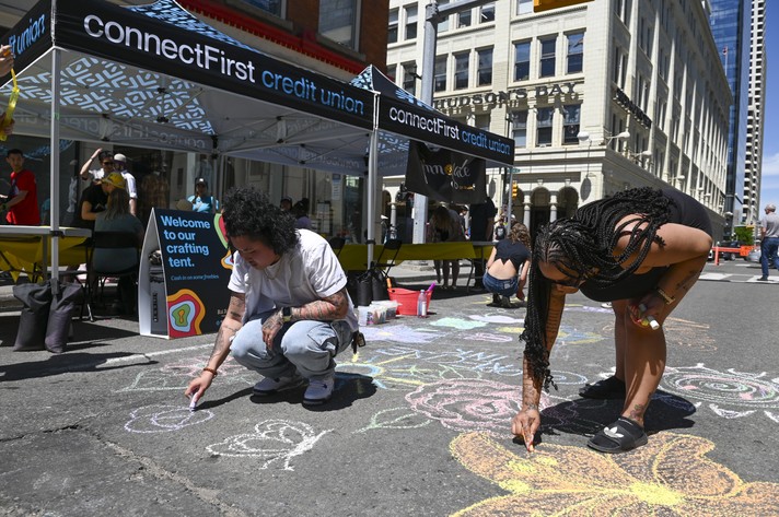 Several people are seen drawing on the street with sidewalk chalk at Sled Alley during Sled Island 2024. Two connectFirst booths with branded tents sit in the background.