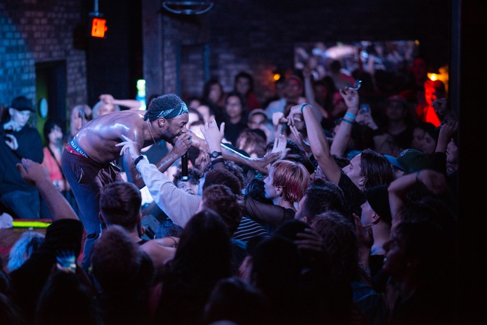 Artist JPEGMAFIA reaches into the crowd while rapping into a microphone during a packed show at Commonwealth Bar & Stage during Sled Island 2019. The rapper is seen shirtless with brown pants and a black bandana headband on.