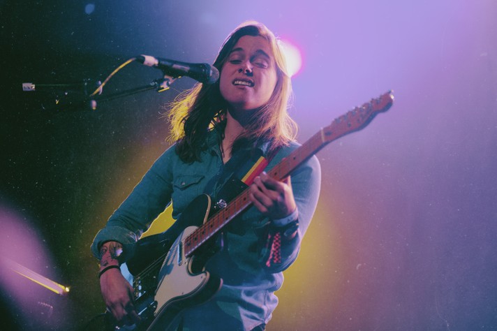 Artist Julien Baker sings and plays guitar at The Palace Theatre during Sled Island 2019. They are wearing a denim shirt, and purple and yellow lights are seen behind them.