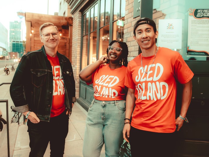 Three volunteers wearing red Sled Island t-shirts smile for the camera outside of the BLOX Arts Centre during Sled Island 2024.