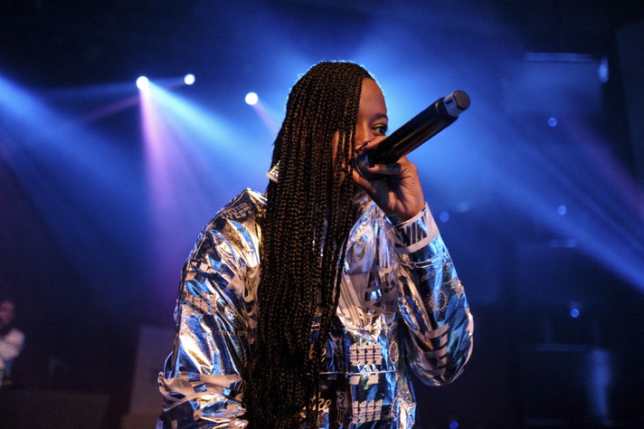 Artist Rapsody raps into a microphone during their performance at The Palace Theatre during Sled Island 2019. They are seen wearing a shiny metallic like jacket and have long braids hanging in front of the right side of their face.