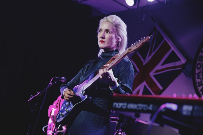 Artist Cate Le Bon is seen playing guitar on stage at the #1 Royal Canadian Legion during Sled Island 2019. They are dressed in all black, with slicked back blonde hair and red lipstick.