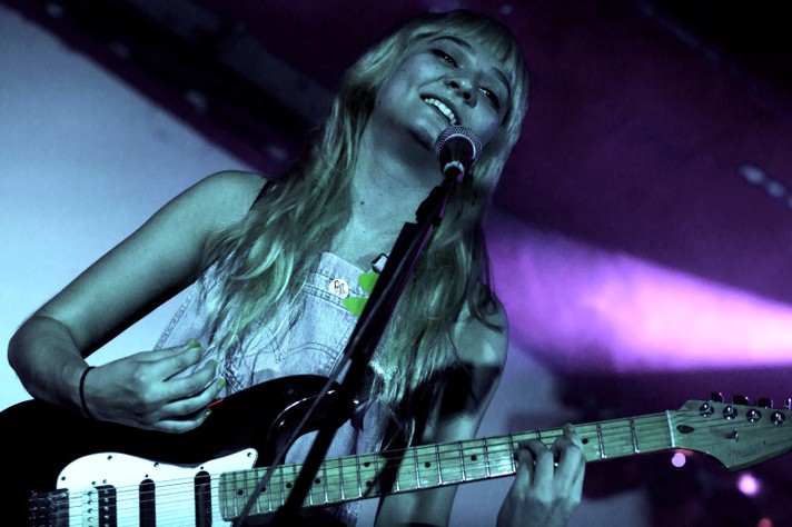 Clementine Creevy of Cherry Glazzer smiles and plays guitar during their performance at the #1 Royal Canadian Legion during Sled Island 2018. Creevy is seen with longe blonde hair and blue overalls.