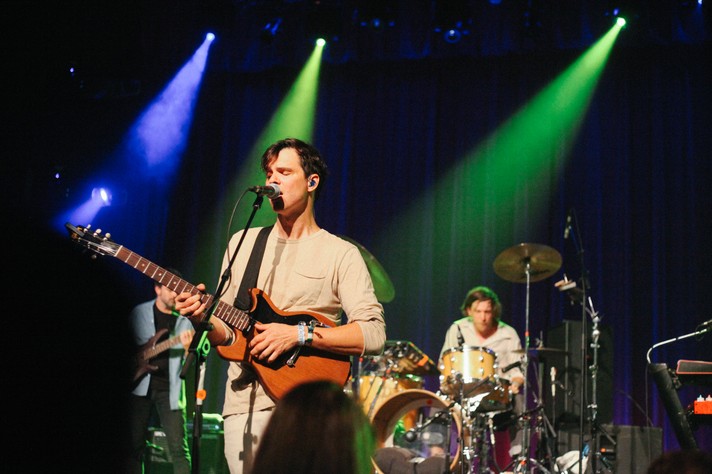 The group Dirty Projectors perform at The Palace Theatre during Sled Island 2018. The front person is shown at the forefront singing into a microphone and playing guitar, while the drummer and the bass player sit in the background. Blue and green lights shine down on them.