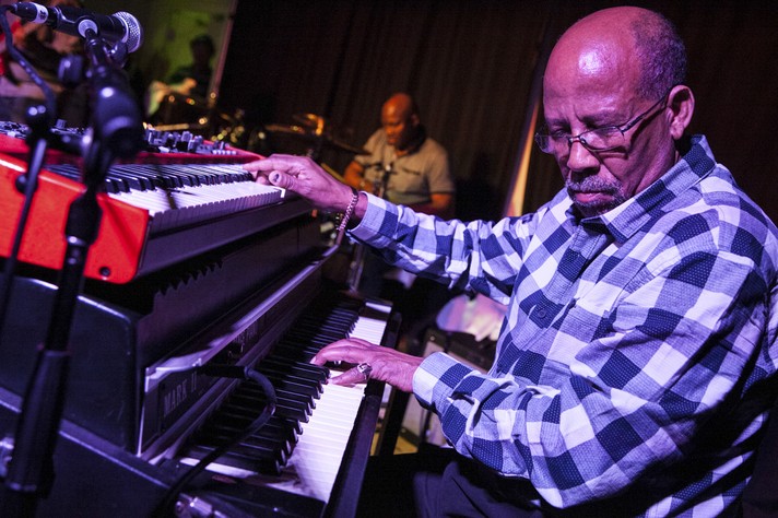 Musician Hailu Mergia is shown playing the organ during a performance at the King Eddy during Sled Island 2017. They are wearing a blue and white checkered shirt and eye glasses.