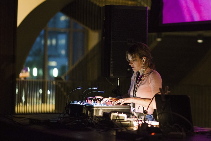 Electronic artist Kaitlyn Aurelia Smith is shown performing at Studio Bell during Sled Island 2017. They are seen with blonde tied up hair, wearing a pink sweater, blue jeans, and colourful dangly earrings.
