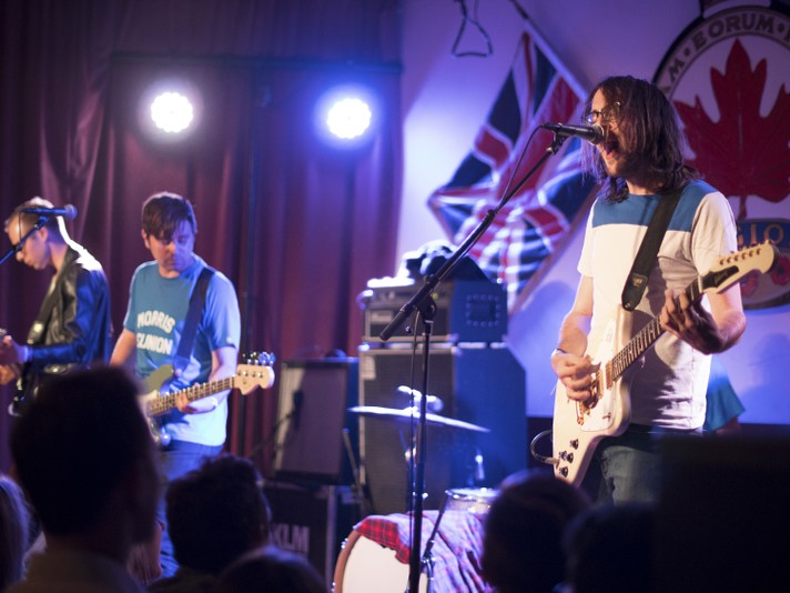 Three members of the group Cloud Nothings are shown performing at the #1 Royal Canadian Legion during Sled Island 2017. The frontperson is shown on the right hand side, playing guitar and singing into the microphone wearing a blue and white t-shirt.