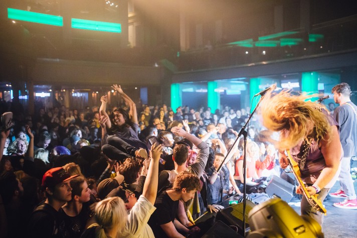 The group Wavves is seen playing on stage at The Palace Theatre during Sled Island 2017. The guitar player in the bottom right of the frame is seen headbanging their long hair. In front of him, an audience member crowd surfs.