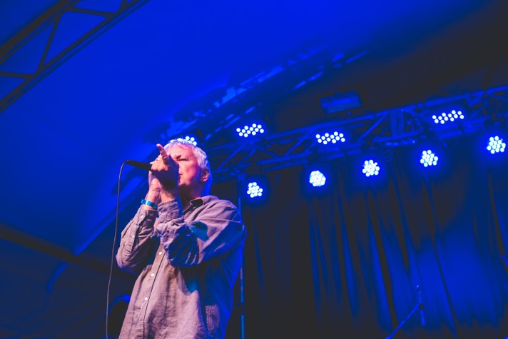 The frontperson of the group Guided by Voices sings into the microphone and points out to the crowd during their performance at Olympic Plaza during Sled Island 2016. Blue stage lights shine in the background.