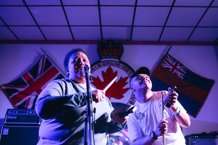 Two members of the group ESG are seen performing at the #1 Royal Canadian Legion during Sled Island 2016. Both people are hitting a cowbell with a drum stick, and the person on the left is seen singing into a microphone.