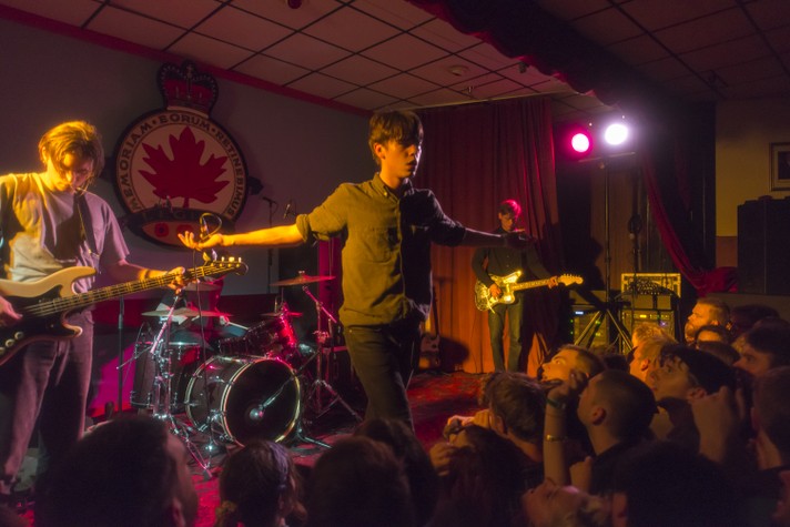 The group Iceage is shown performing at the #1 Royal Canadian Legion during Sled Island 2013. The frontperson is at the centre of the stage with their arms spread out in a cross formation.