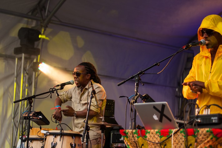 Two members of the group Shabazz Palaces are seen performing at Olympic Plaza during Sled Island 2012. One sits behind a conga set, and the other behind a DJ set up.