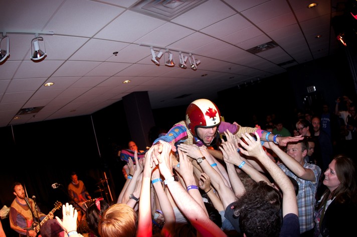 Nardwuar performs as part of the group The Evaporators at National Music Centre during Sled Island 2012. He crowd surfs wearing a helmet with a red maple leaf on it while the band plays on the stage behind him.