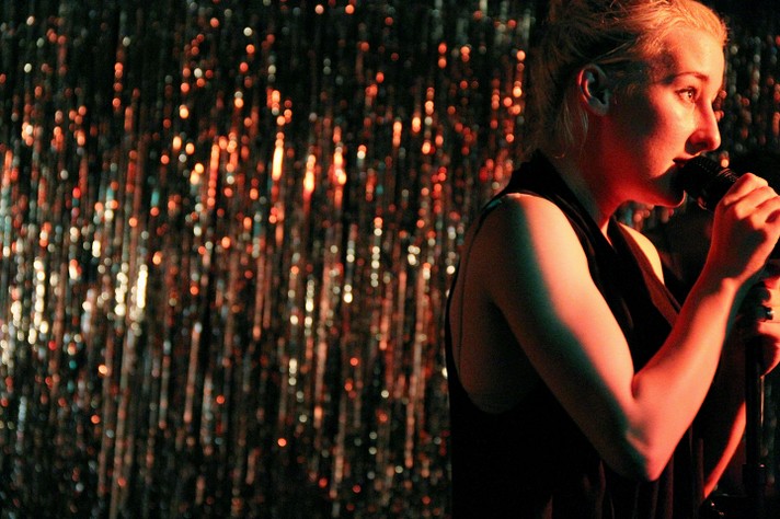 The frontperson of Zola Jesus is seen with short blonde hair and a sleeveless shirt singing into a microphone in front of a shimmering silver background at The Marquee Room during Sled Island 2011.