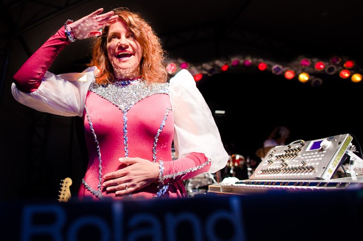 A member of the group Of Montreal is shown wearing a pink and white jumpsuit and long red hair smiling and holding their hand up to their head at Olympic Plaza during Sled Island 2011.