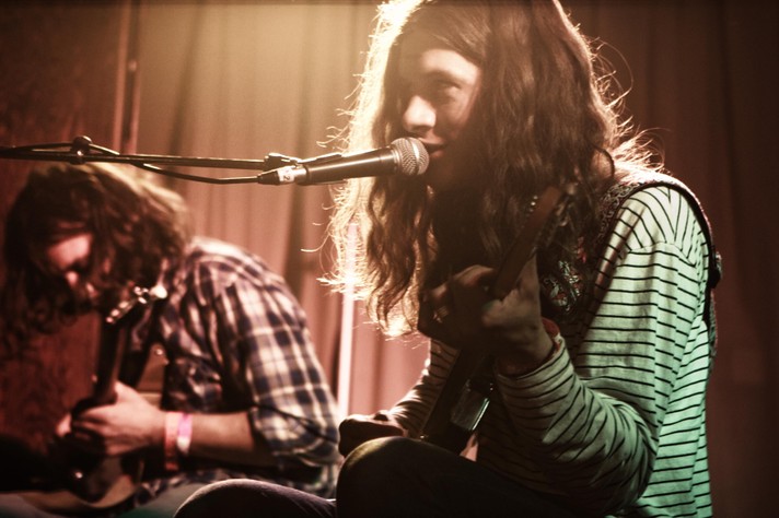 Artist Kurt Vile is shown sitting down playing guitar and singing into a microphone at Hillhurst United Church during Sled Island 2011. He has a green and black striped long sleeve t-shirt on, and long dark hair. A second guitar player is shown to his left.