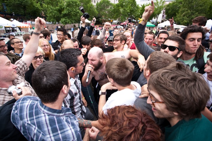 The singer of the group Fucked Up is shown in the middle of the audience, singing in the microphone while shirtless during their performance at Olympic Plaza at Sled Island 2010.