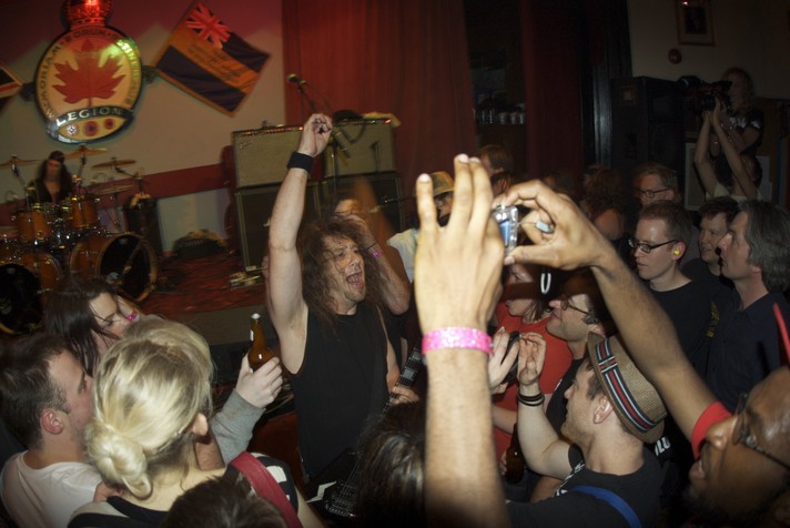 A member of the group Anvil is shown standing in the crowd with his guitar and his arm in the air during their performance at the #1 Royal Canadian Legion during Sled Island 2009.