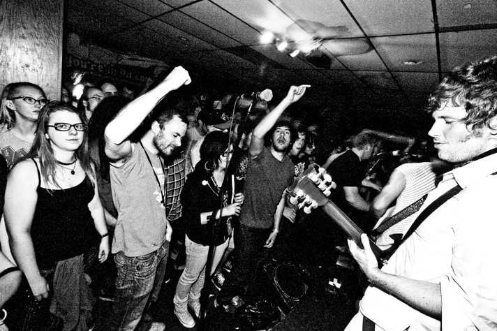 A black and white photo shows the band Tricky Woo performing in front of an audience at Verns during Sled Island 2009. Several audience member have their arms in the air.