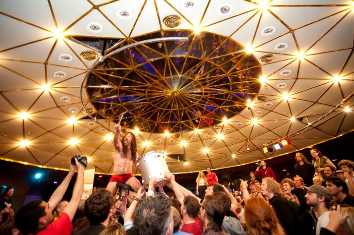 A member of the group Monotonix is lifted up by the audience and along with a drum during their performance at The Distillery for Sled Island 2009. The circular ceiling is seen above them, which has bright lights, elements of silver and gold, and a mirrored centre.