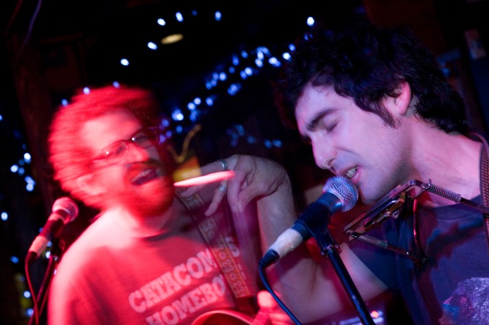 Two members of the group Blitzen Trapper are shown performing at Broken City during Sled Island 2008. The guitar player is on the right bathed in red light and slightly out of focus, and the other member is on the right and is shown singing into the microphone with their eyes closed.