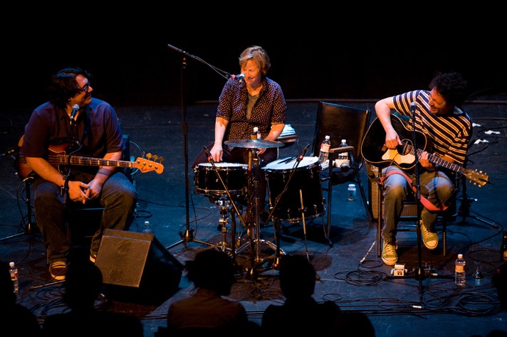 The three members of the group Yo La Tengo perform while sitting at the Pumphouse Theatre for Sled Island 2008. The members on the left and right hold guitars, while the one in the middle has a small drum kit.