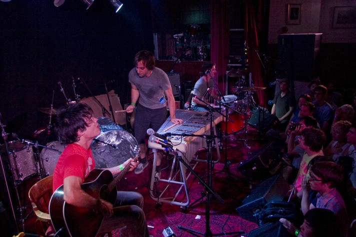 The group The Dodos are shown performing at the #1 Royal Canadian Legion during Sled Island 2008. The drummer and acoustic guitar player are seated on the left and right sides. The member in the middle appears to be playing both a xylophone and a trash can.