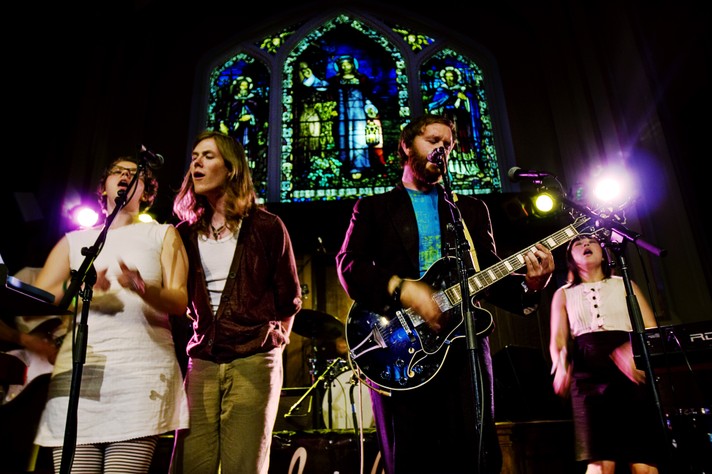 Four members of the group Woodpigeon are seen singing in unison during their performance at Central United Church for Sled Island 2008. A religious stain glass window is in the background.