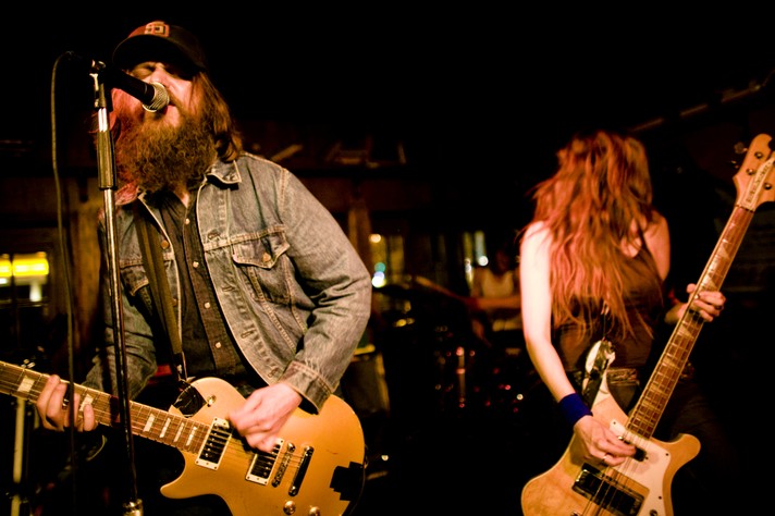 Two members of the group C'mon are shown performing at the Ship & Anchor Pub during Sled Island 2007. The guitar player on the left is seen singing wearing a jean jacket and black ball cap. The bass player on the right has long dark hair and is wearing a black tank top.