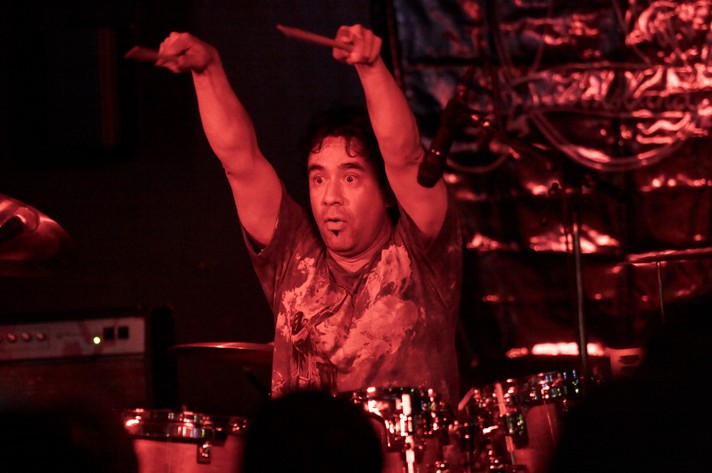 Comedian and musician Fred Armisen is shown behind a drum kit with his arms in the air pointing the drum sticks toward the audience during his performance at The Warehouse for Sled Island 2007.