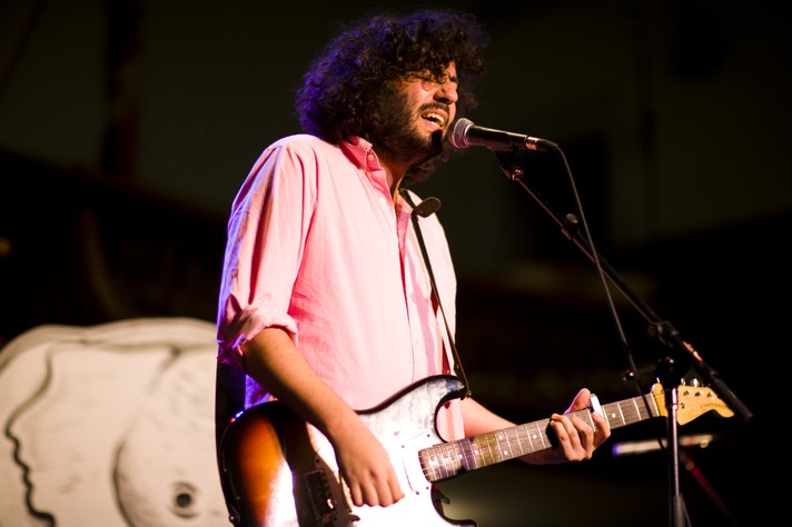 The frontperson for the group Destroyer is seen singing and playing guitar during their performance at Grace United Church for Sled Island 2007. They have long dark curly hair and a beard, and are wearing a pink button up shirt.