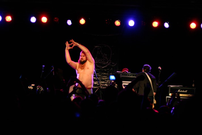 The frontperson of the group Les Savy Fav is seen shirtless holding their arms in air and making a heart shape with their hands during their performance at MacEwan Ballroom for Sled Island 2007.