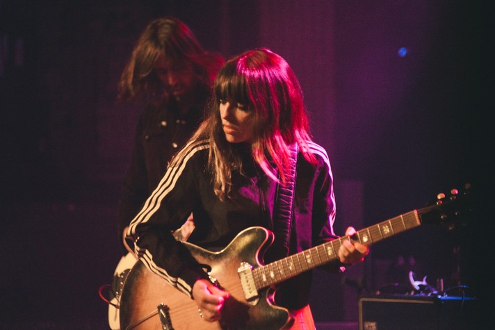 The lead singer of the group Hop Along is shown playing guitar at The Palace Theatre during Sled Island 2019. They have long brown hair and black sport coat.