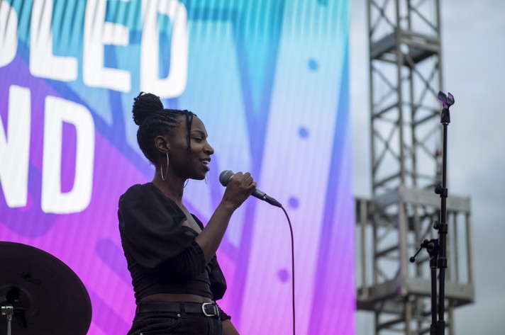Artist Indigo Rose performs at at High Park for Camp Sled Island in 2021. They are seen singing into a microphone in front of a colourful screen that says "Welcome to Sled Island."