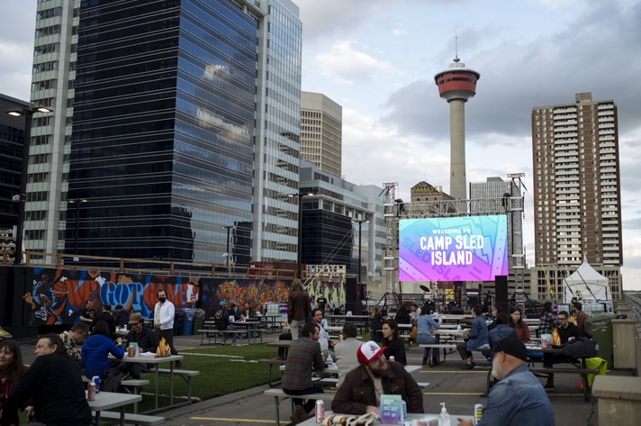 A photo of Camp Sled Island - an event that took place at High Park. People are sitting at picnic tables that set up on an open rooftop, and a large video screen sits at the end that says "Camp Sled Island." The Calgary Tower and other tall buildings are seen in the background.