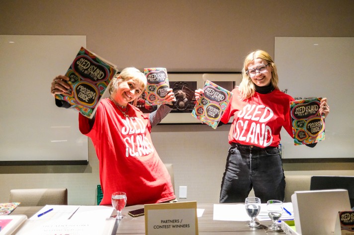 Two volunteers in red Sled Island shirts hold up the 2024 program guide at Pass Pick-Up at the Delta Hotel
