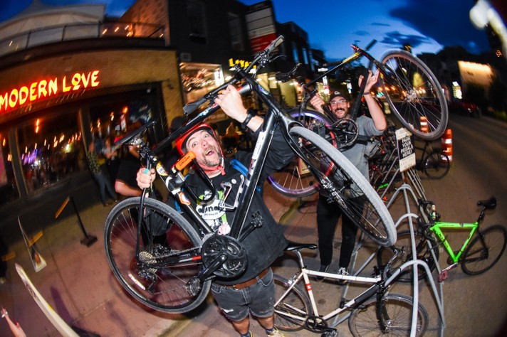 Two attendees lift their bikes over their heads for a photo at the bike racks set up outside of Modern Love during Sled Island 2024.