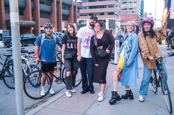 Six festival attendees smile for a photo outside of Modern Love during Sled Island 2024. The people on the left and right side are on bikes, while the others are on foot.