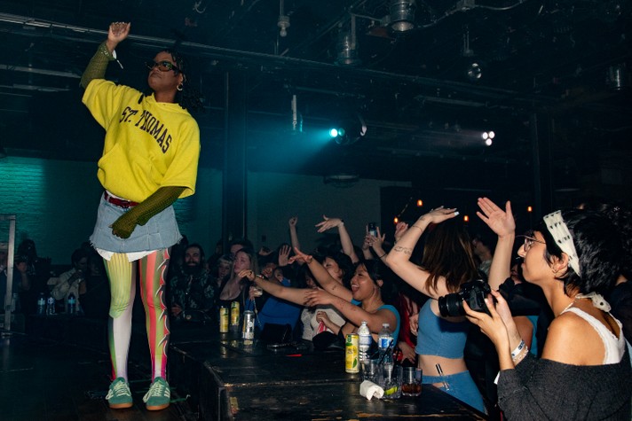 Artist Kari Faux is seen on stage wearing a yellow hoodie, denim skirt and colourful leggings during their performance at Commonwealth Bar & Stage during Sled Island 2024.